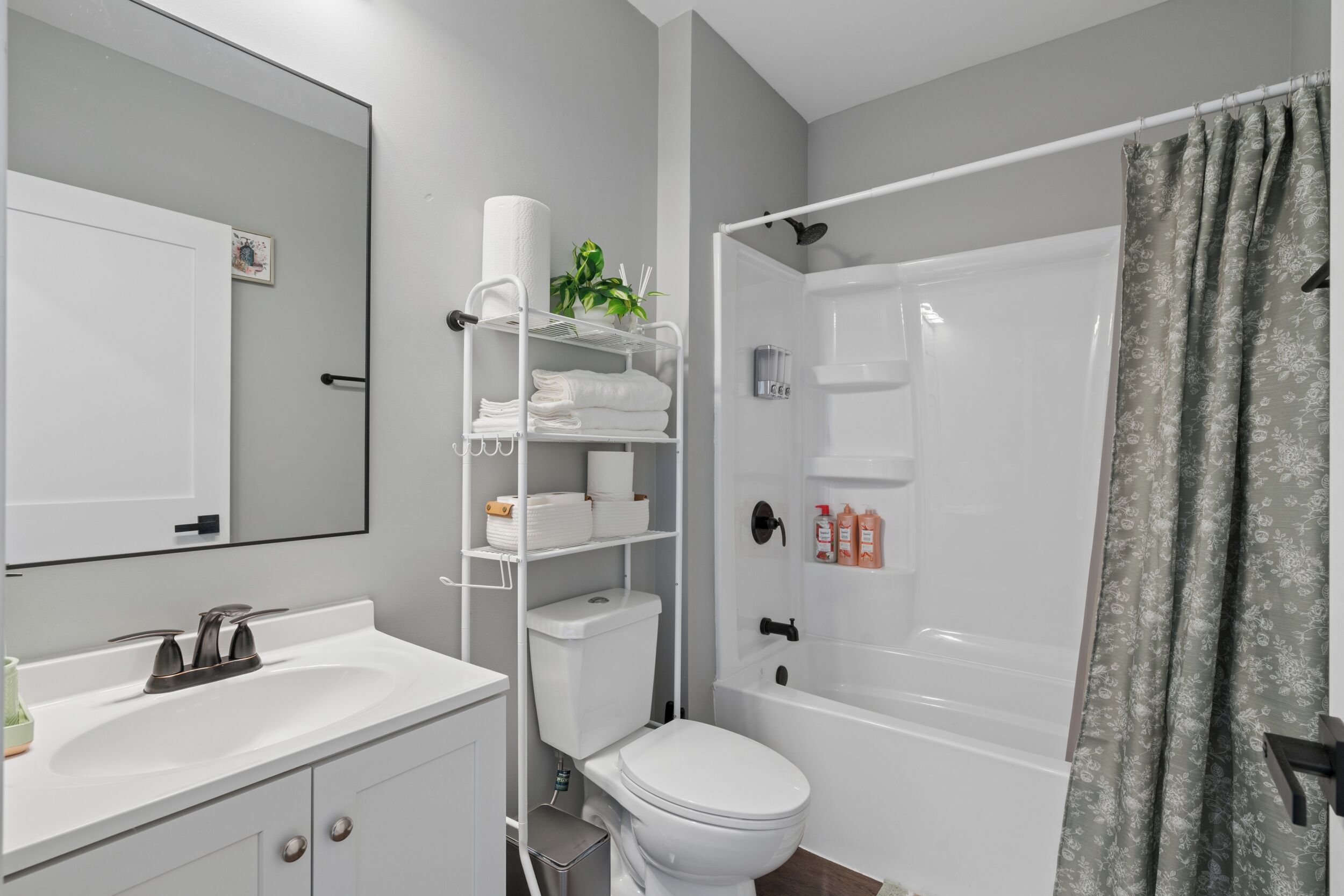 Un bagno moderno con lavandino, water e vasca da bagno bianchi. Si nota uno scaffale con articoli da toilette e una pianta.