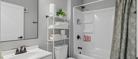 Un bagno moderno con lavandino, water e vasca da bagno bianchi. Si nota uno scaffale con articoli da toilette e una pianta.