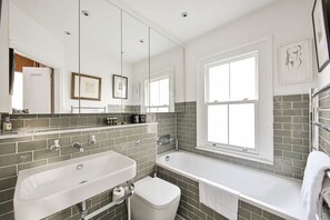 A modern bathroom with a large bathtub, a separate sink, and a toilet. There are two windows providing natural light. - Argyl | Bright 3 Bedroom Home in Shepherd’s Bush (Greater London)
