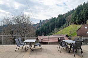 A terrace with outdoor seating, a view of a mountainous landscape, and a building with a red-tiled roof. - Apartment 5 with Balcony, Barbecue Area and Wi-Fi (Oberharmersbach)