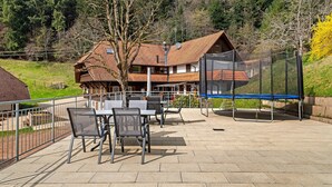 Une terrasse avec des sièges d’extérieur, un trampoline et une maison avec un toit en pente, le tout situé dans une zone boisée.