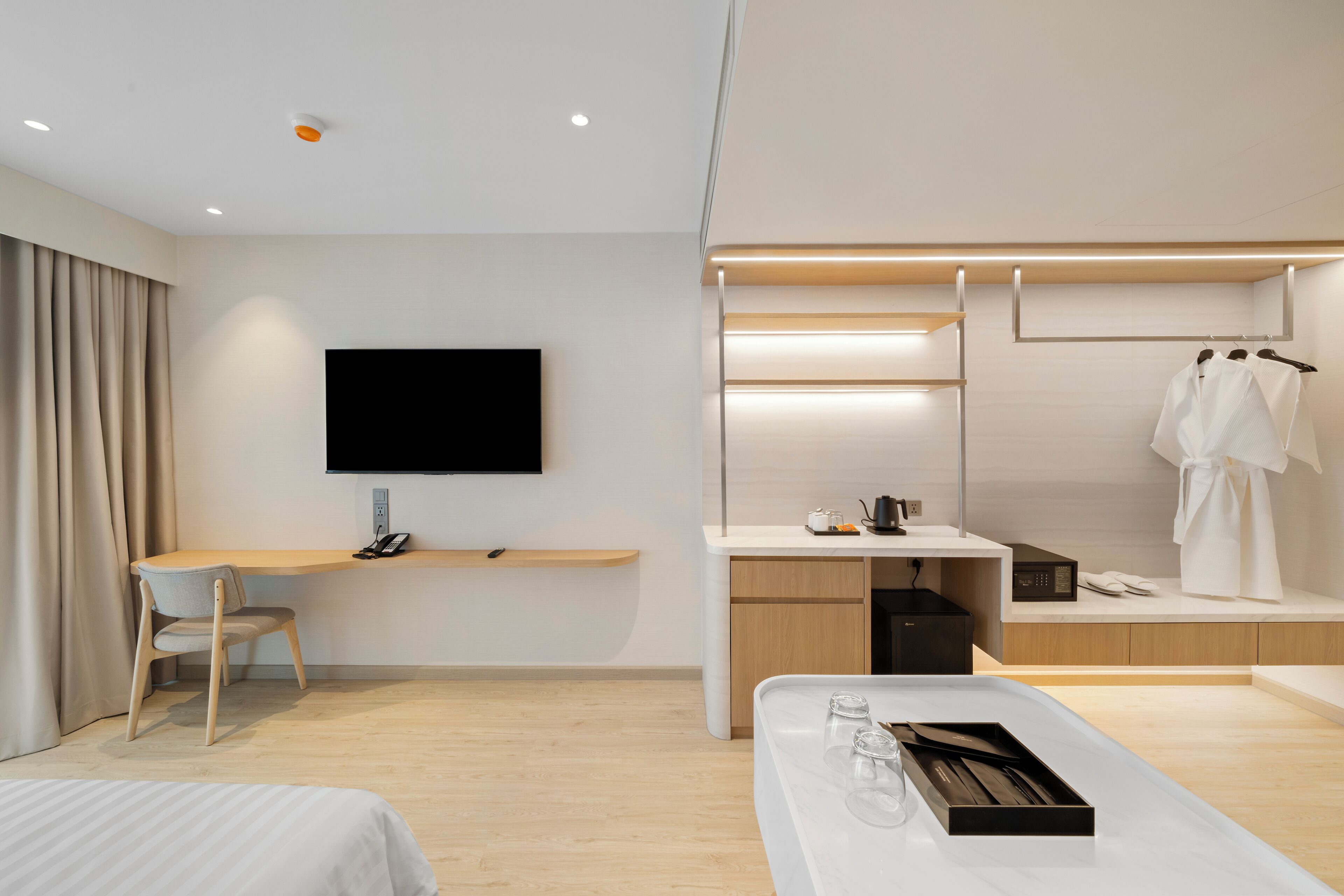 A modern hotel room with a flat-screen TV, a desk with a chair, a bed, a bedside table with glasses and a tray, and a wardrobe with clothes.