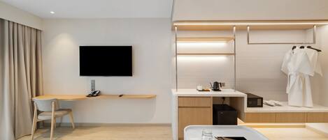 A modern hotel room with a flat-screen TV, a desk with a chair, a bed, a bedside table with glasses and a tray, and a wardrobe with clothes.