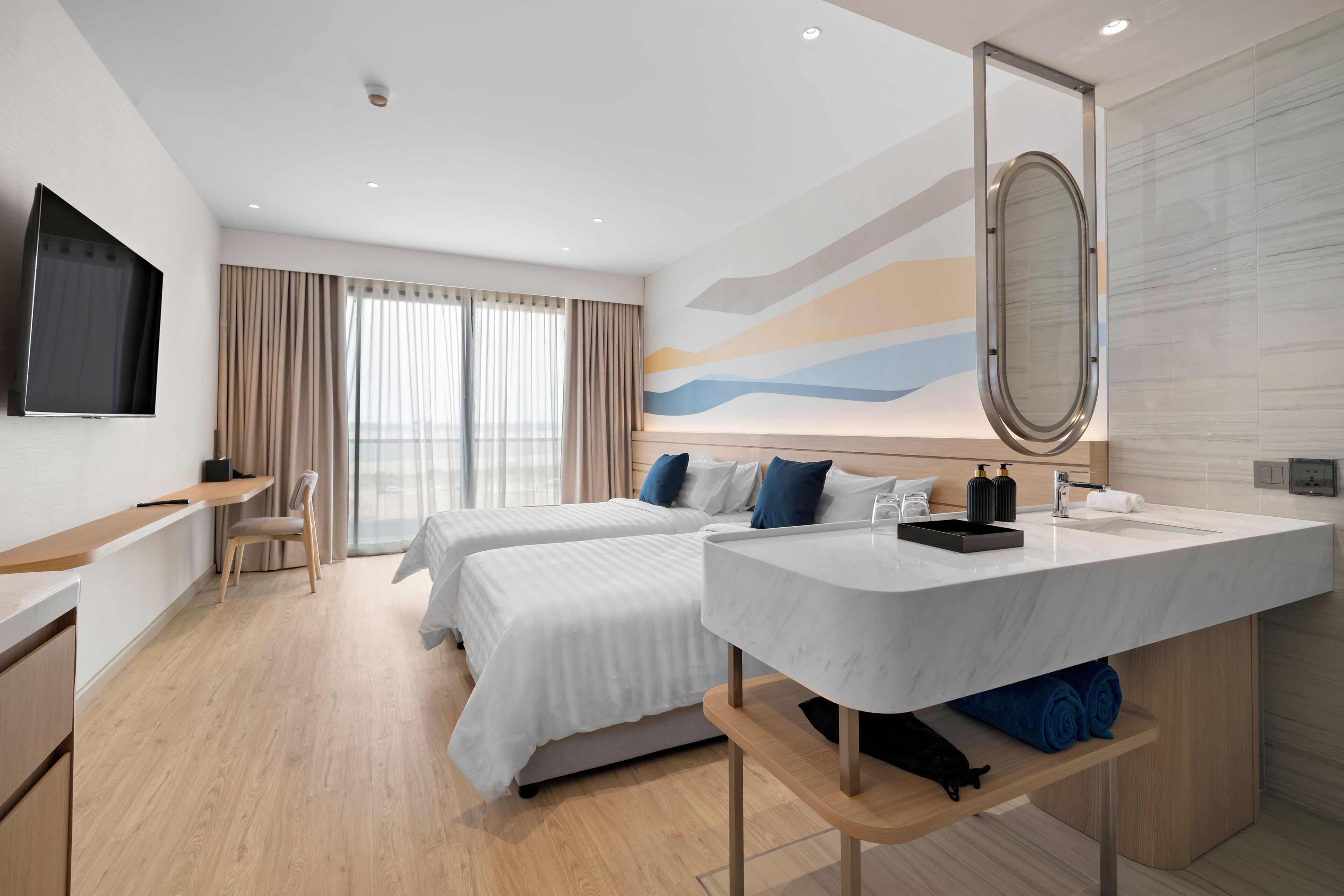 A hotel room with two beds, a desk, a TV, a bathroom with a mirror, and a large window.