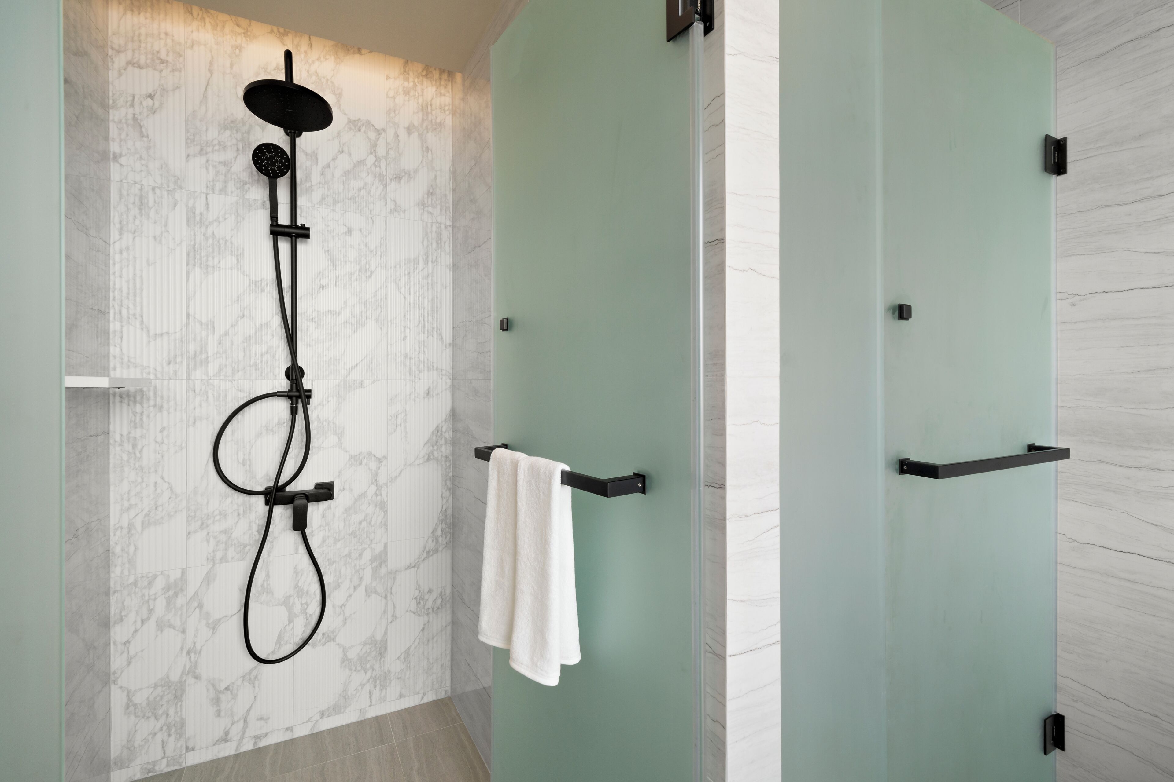 Un baño moderno con pared de mármol, ducha con manguera negra y un perchero con toalla blanca.