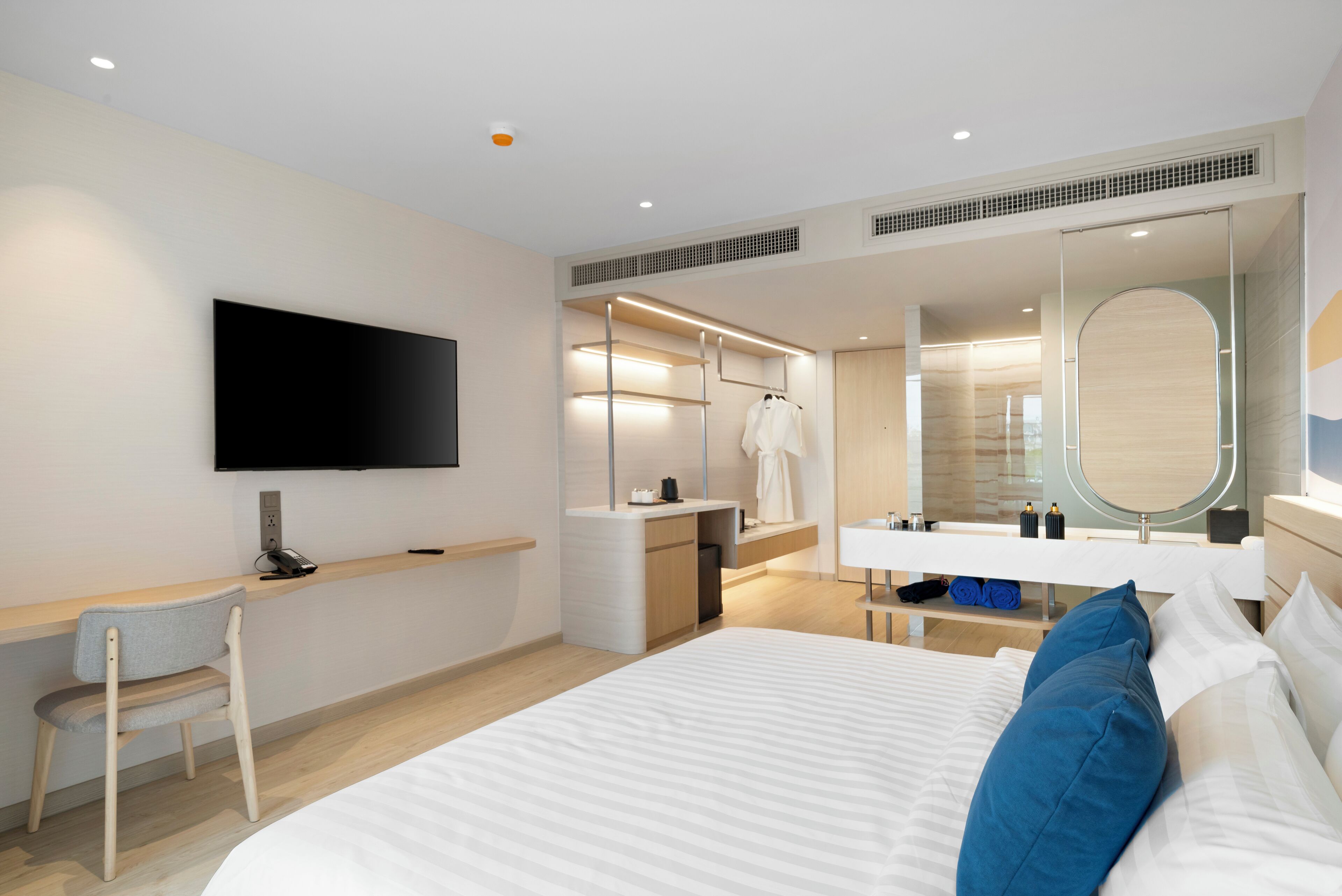 A modern hotel room with a bed, a desk, a chair, a TV, a wardrobe, and a bathroom with a mirror.