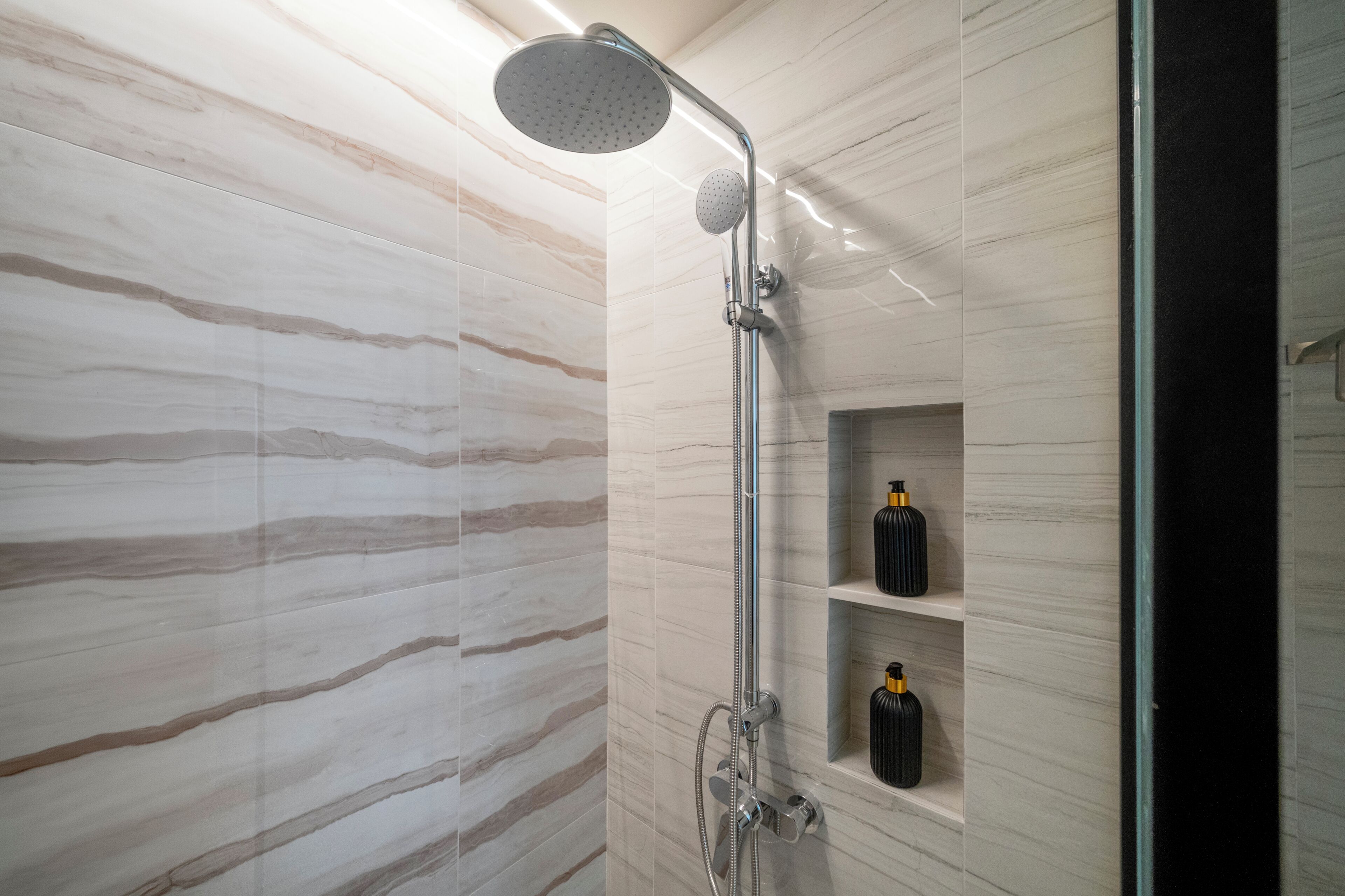 Ducha moderna con cabezal de ducha de lluvia y cabezal de ducha de mano, paredes de mármol y repisa con botellas.