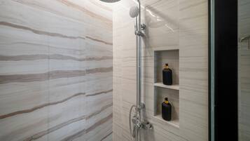 Ducha moderna con cabezal de ducha de lluvia y cabezal de ducha de mano, paredes de mármol y repisa con botellas.