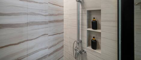 A modern shower with a rain showerhead and handheld showerhead, marble walls, and a shelf with bottles.