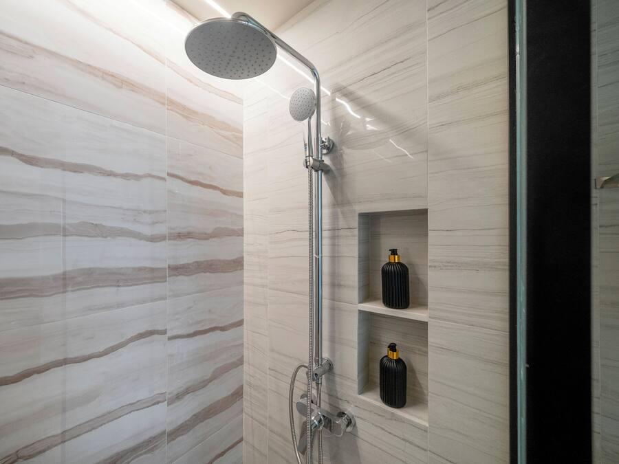 Ducha moderna con cabezal de ducha de lluvia y cabezal de ducha de mano, paredes de mármol y repisa con botellas.