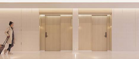 A modern hotel lobby with a clean, minimalist design, featuring a central corridor with doors on either side and a person walking with a suitcase.