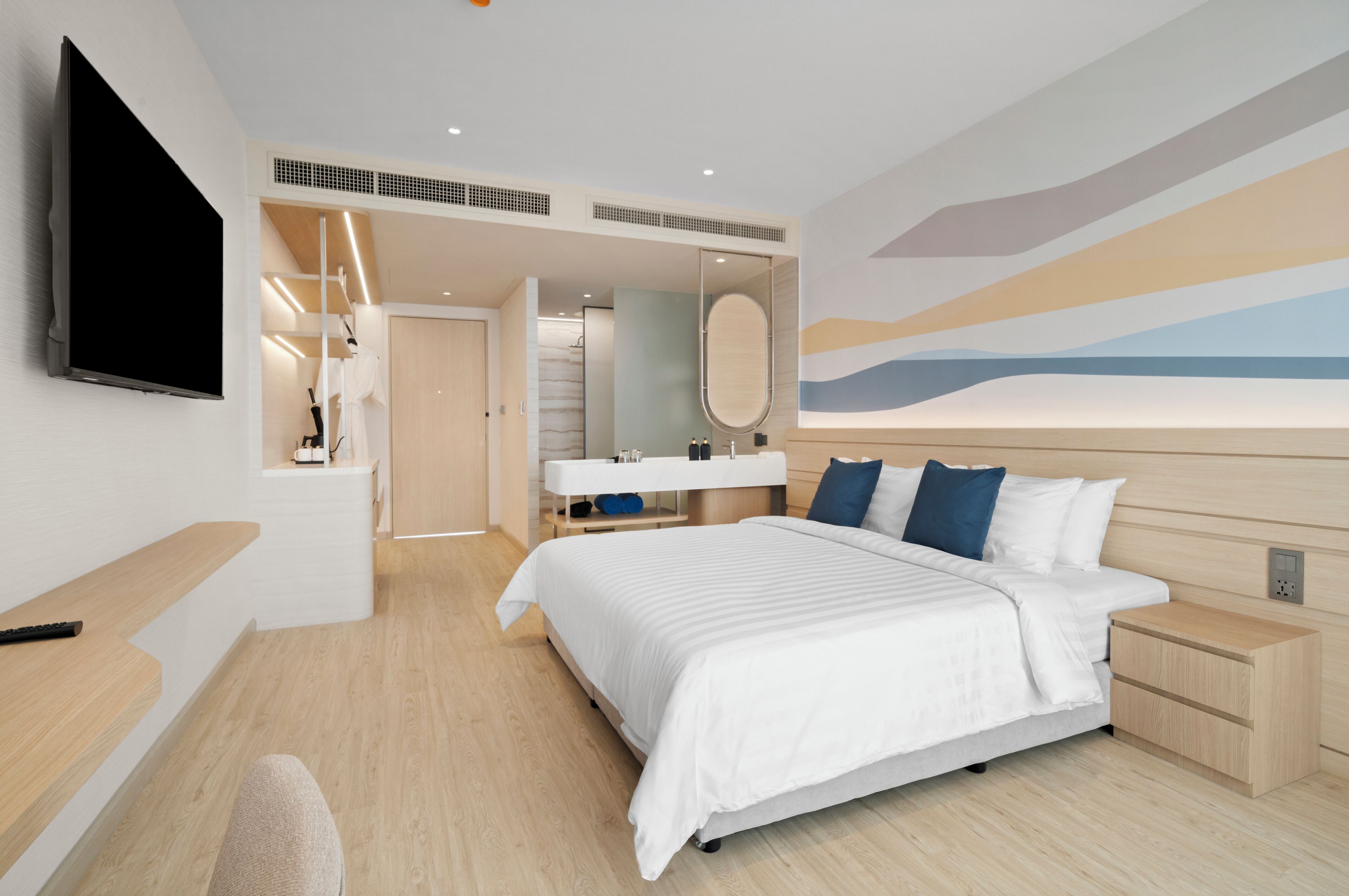 A modern hotel room with a large bed, a TV mounted on the wall, a vanity with a mirror, and a bathroom visible through an open door.