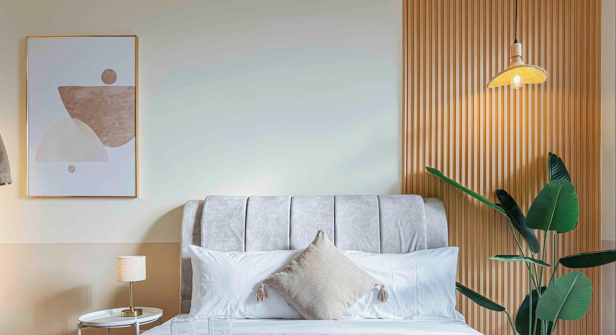 Un dormitorio ordenado con cama, almohadas, una mesita de noche, una lámpara, una obra de arte en la pared y un panel de madera vertical con una luminaria colgante.