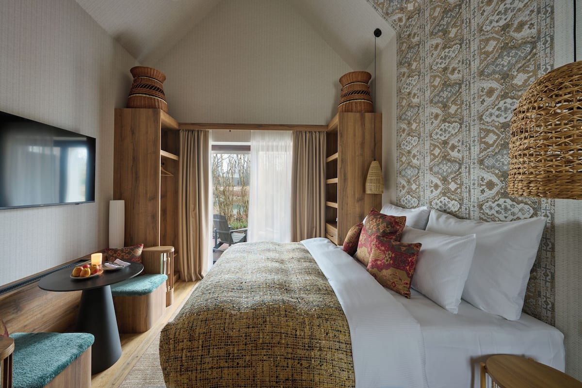 A spacious bedroom with a large bed, a TV mounted on the wall, and a view of the outdoors through a large window.