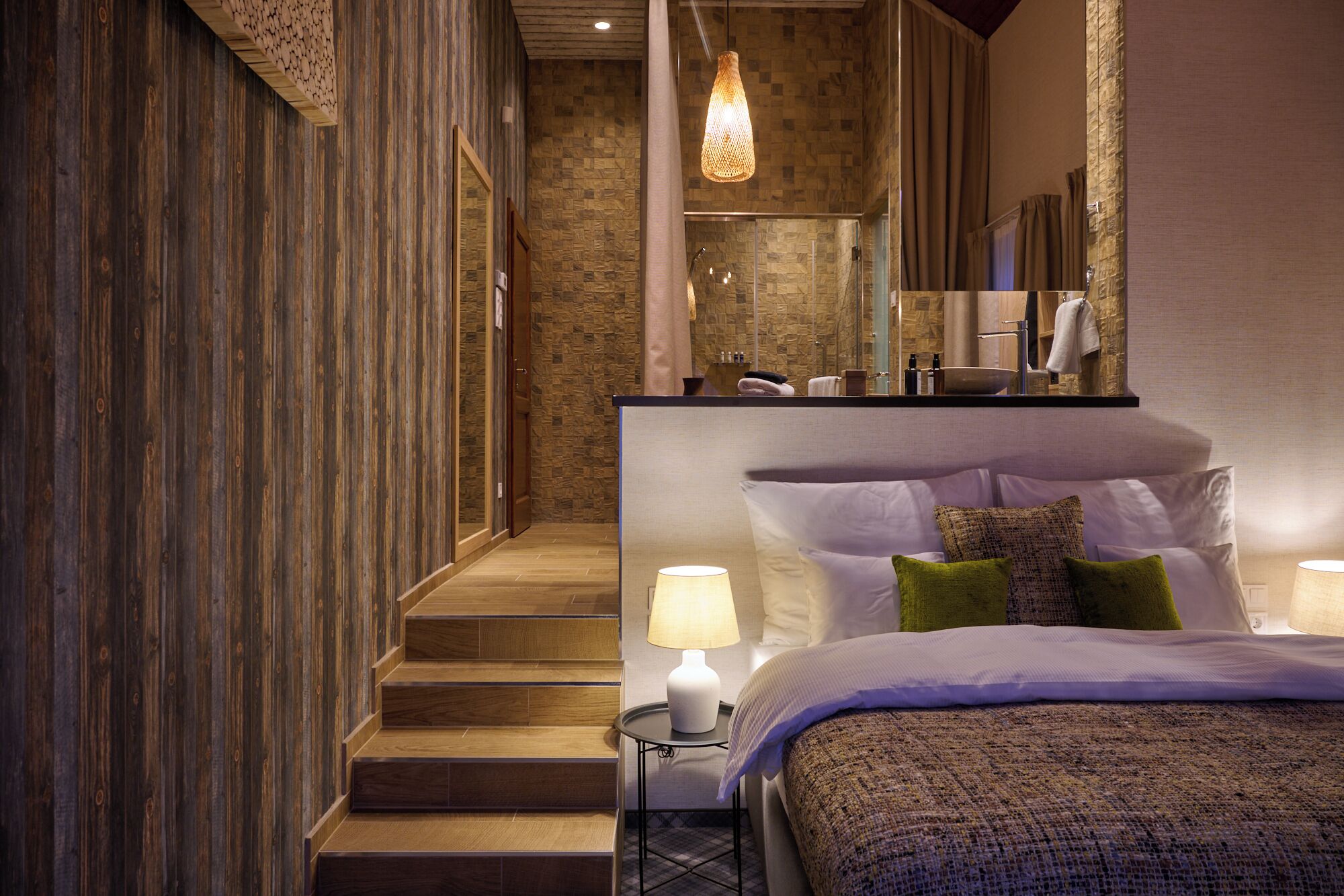 A hotel room with a wooden staircase, a large bed with multiple pillows, a nightstand with a lamp, and a bathroom visible through a glass door.