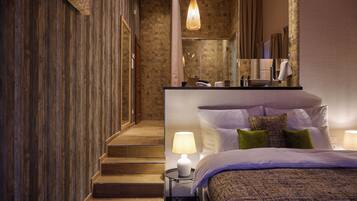 A hotel room with a wooden staircase, a large bed with multiple pillows, a nightstand with a lamp, and a bathroom visible through a glass door.