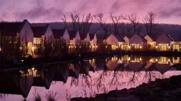 Filas de casas modernas con un lago tranquilo que refleja sus luces, rodeado de árboles y un cielo púrpura.