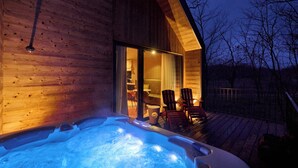 A wooden cabin with a hot tub on a deck, chairs, and a glass door leading inside.