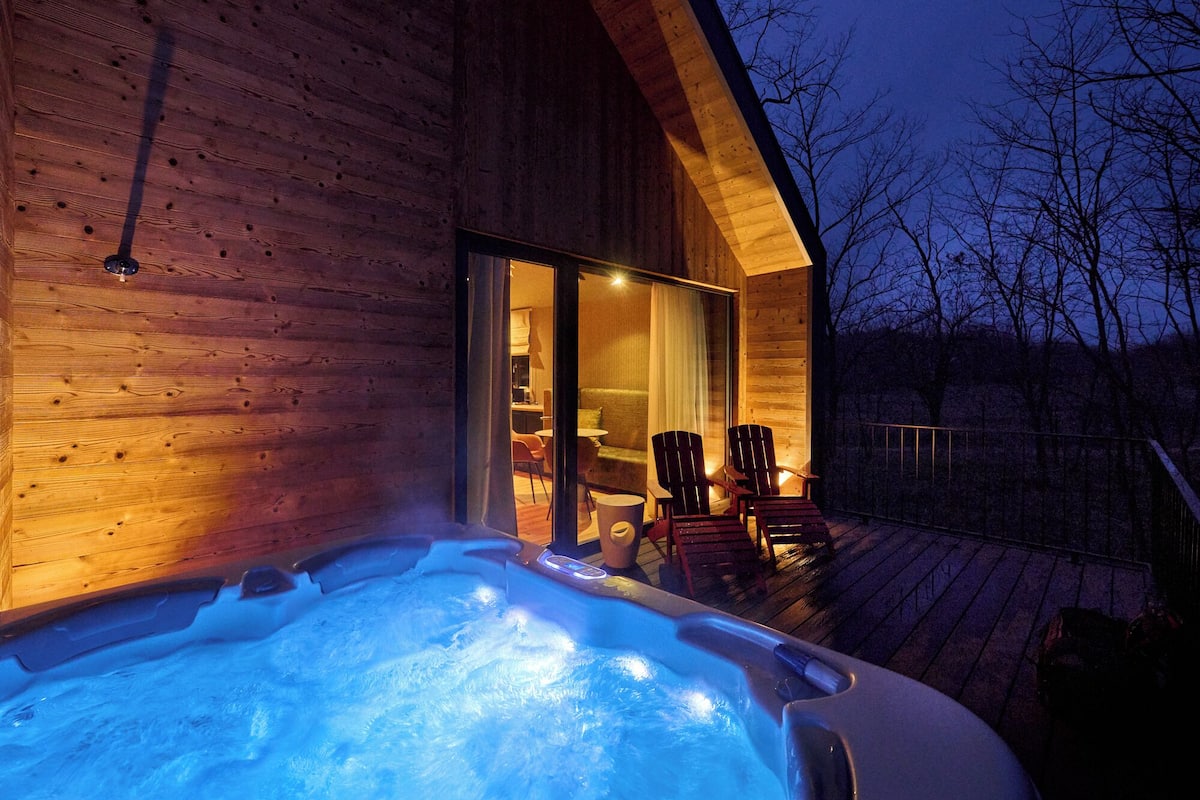 A wooden cabin with a hot tub on a deck, chairs, and a glass door leading inside.