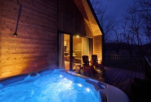 A wooden cabin with a hot tub on a deck, chairs, and a glass door leading inside.