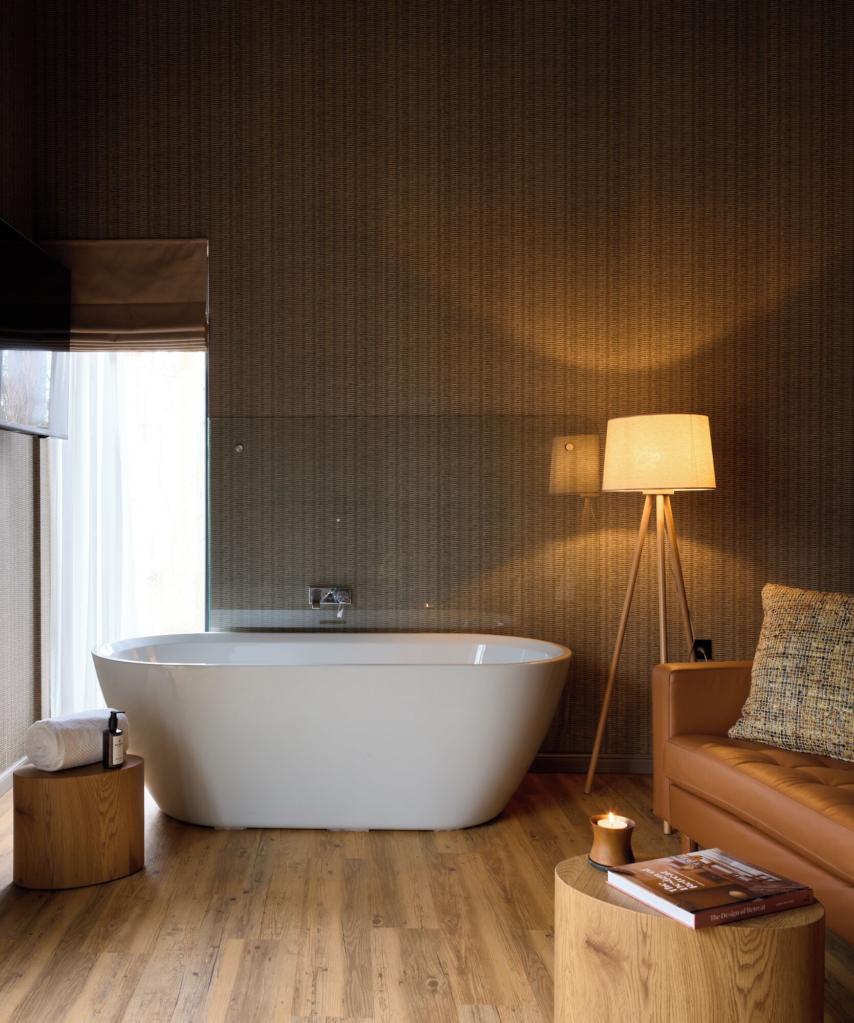 Une salle de bain moderne avec une baignoire sur pieds, une petite table en bois avec un livre, une lampe sur pied et un canapé agrémenté d’un plaid à motifs.