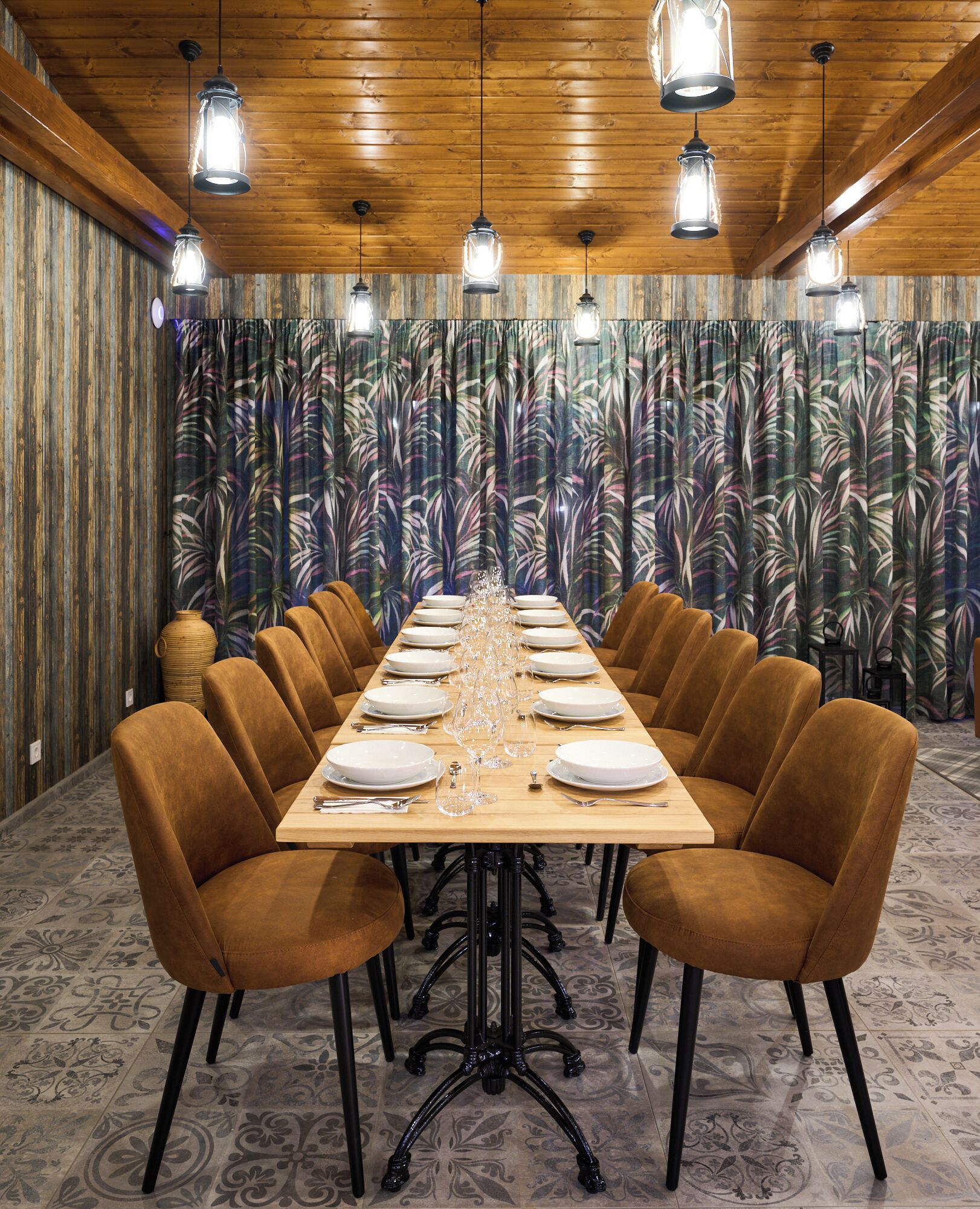 Une longue table à manger, dressée pour un repas, avec des chaises à revêtement marron, un plafond en bois et un rideau décoré de motifs de feuilles tropicales en arrière-plan.