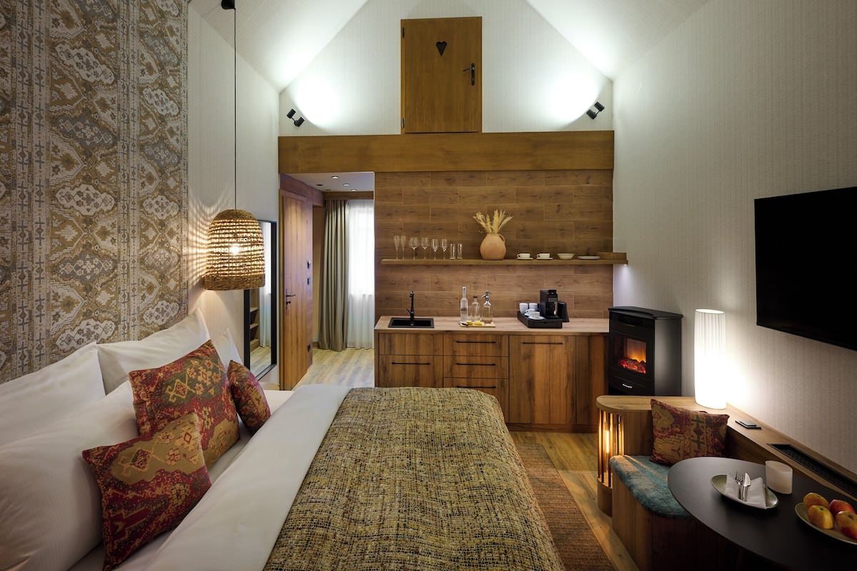 A bedroom with a large bed, a wooden dresser, a small kitchen area with a stove, and a TV mounted on the wall.