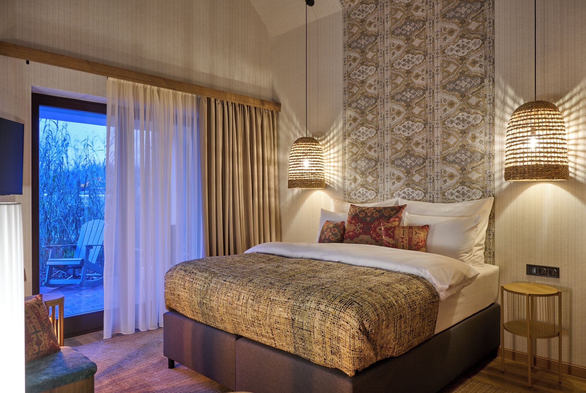 A bedroom with a large bed, two wall-mounted lamps, a patterned headboard, and a view of the outdoors through a sliding door.