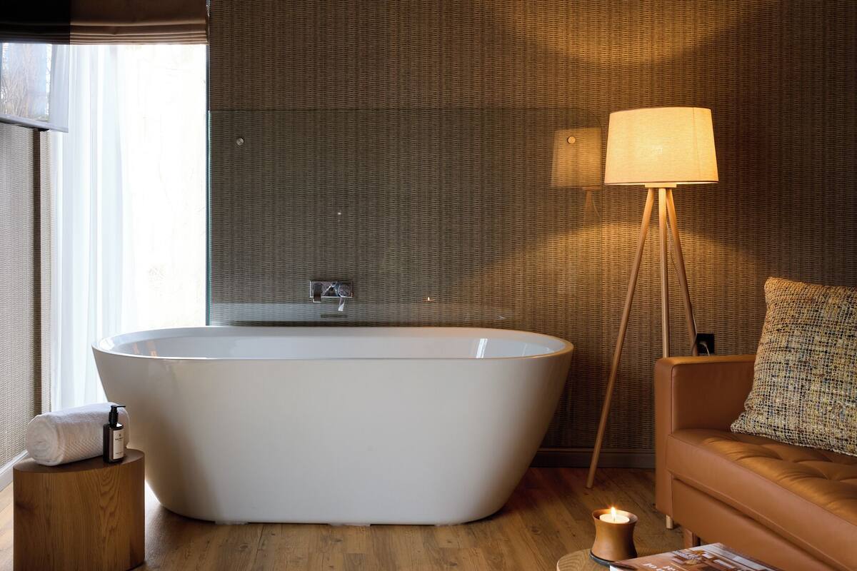 A modern bathroom with a freestanding bathtub, a wooden side table with a book, a floor lamp, and a sofa with a patterned throw.