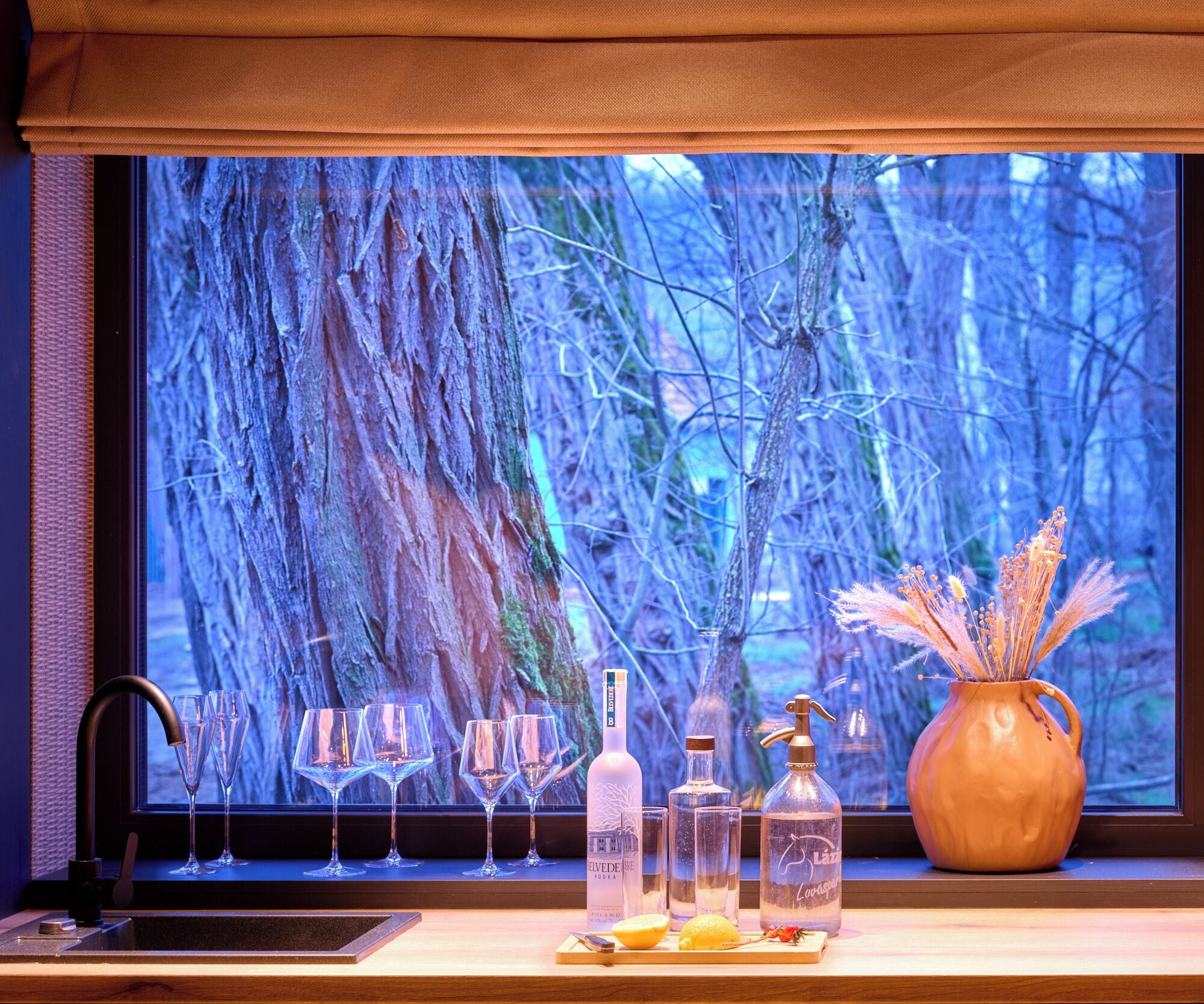Eine Bar mit Blick auf einen Wald durch ein Fenster, mit einer Spirituosenflasche, Gläsern und einer Vase mit Trockenpflanzen.