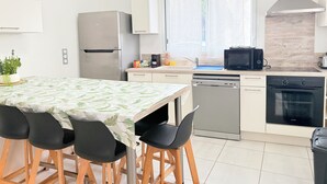 Ett modernt kök med ett kylskåp i rostfritt stål, en mikrovågsugn, en diskmaskin och en spis. Det finns ett matbord med stolar och ett fönster med tunna, genomskinliga gardiner.