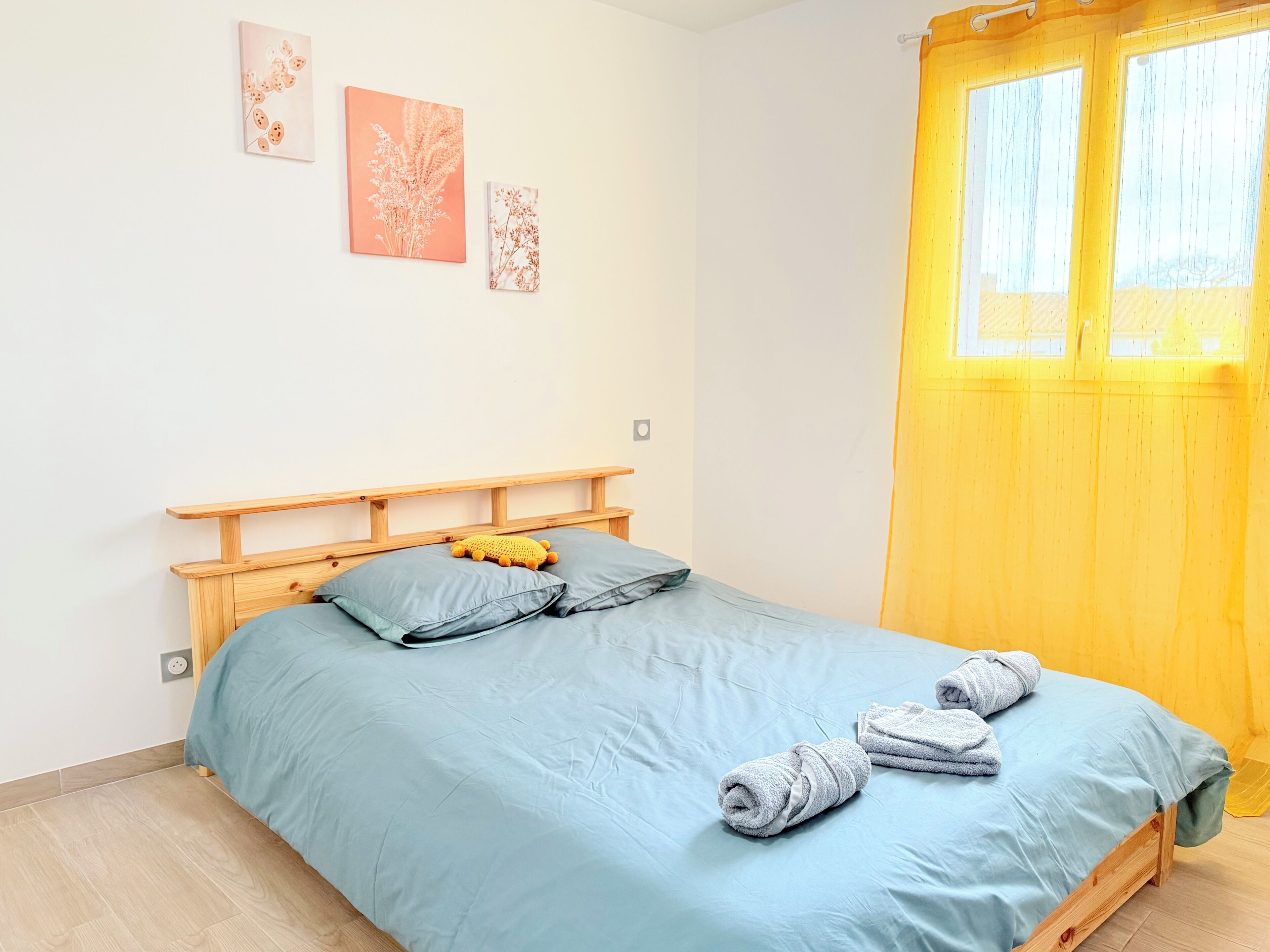 A bedroom with a bed, a wooden headboard, a blue bedspread, two pillows, a yellow curtain, and two wall-mounted artworks.