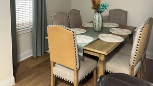 Un coin repas comprenant une table en bois, six chaises rembourrées et un vase avec des fleurs séchées.