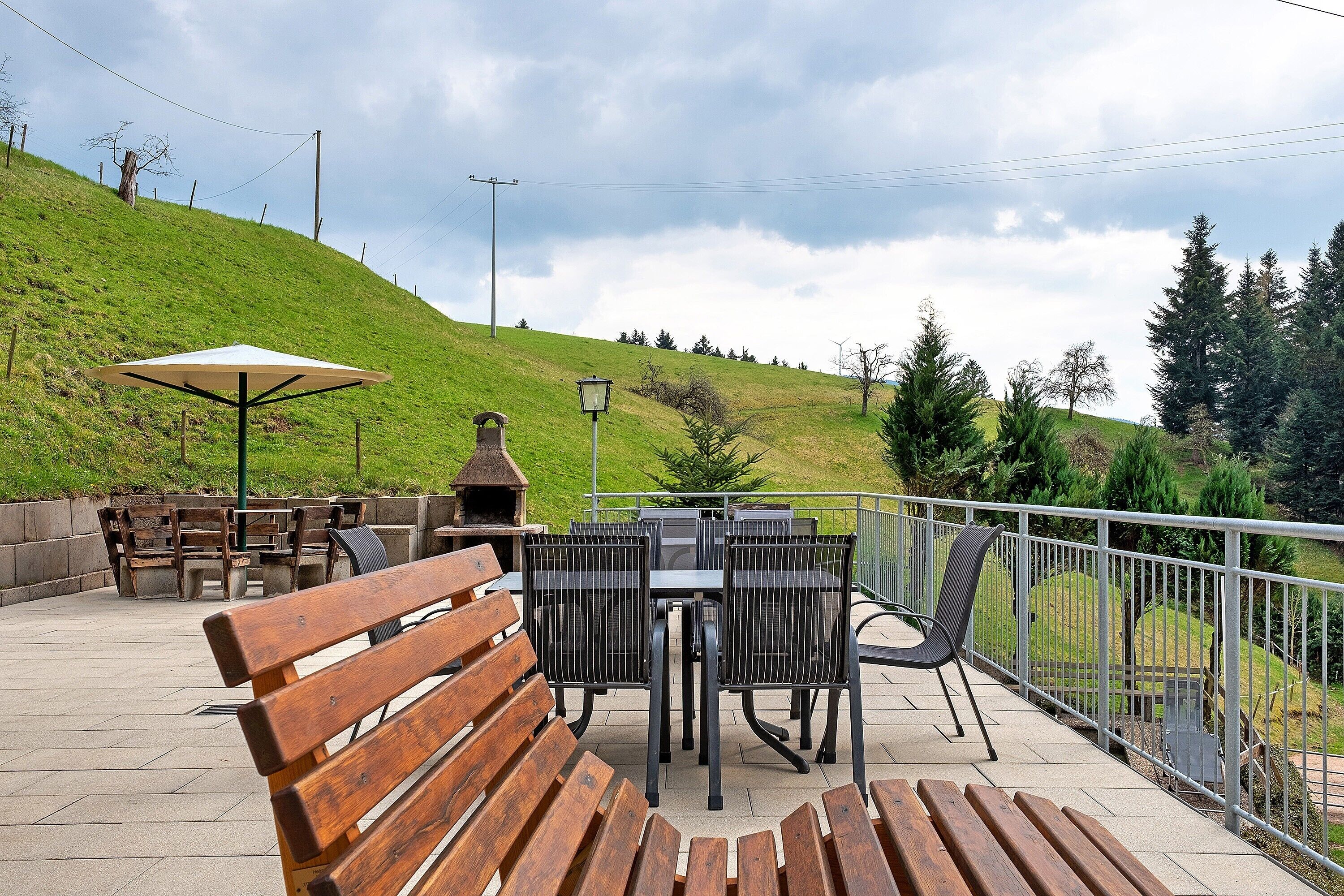 An outdoor patio with wooden benches, a table with chairs, a lamp post, and a view of a grassy hill.