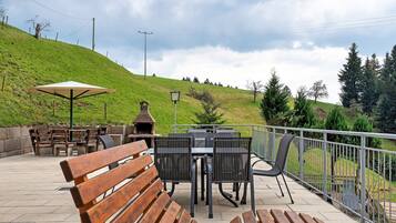 An outdoor patio with wooden benches, a table with chairs, a lamp post, and a view of a grassy hill.