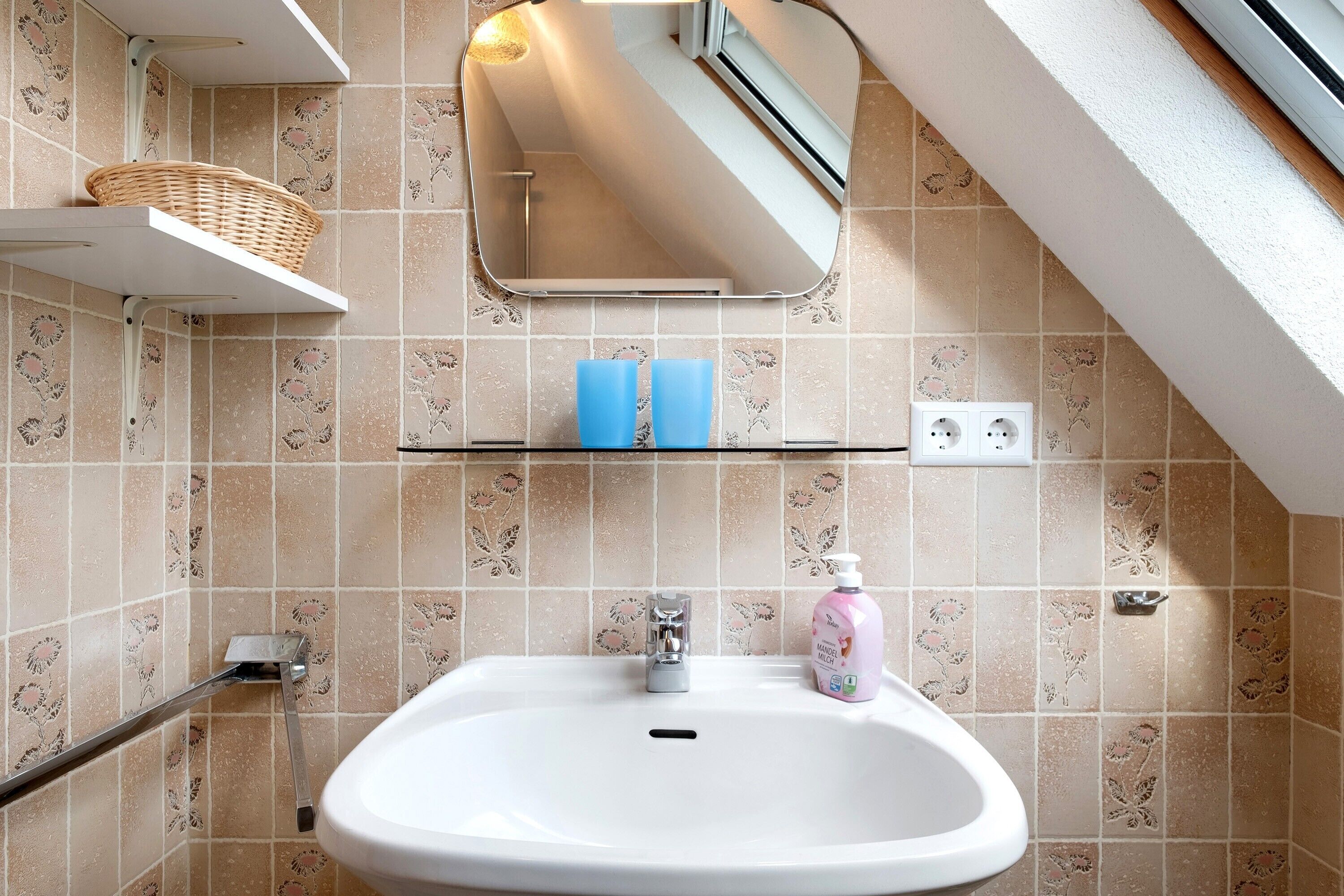 Un bagno con un lavandino bianco, uno specchio, uno scaffale con articoli da toilette e un cesto su uno scaffale bianco.