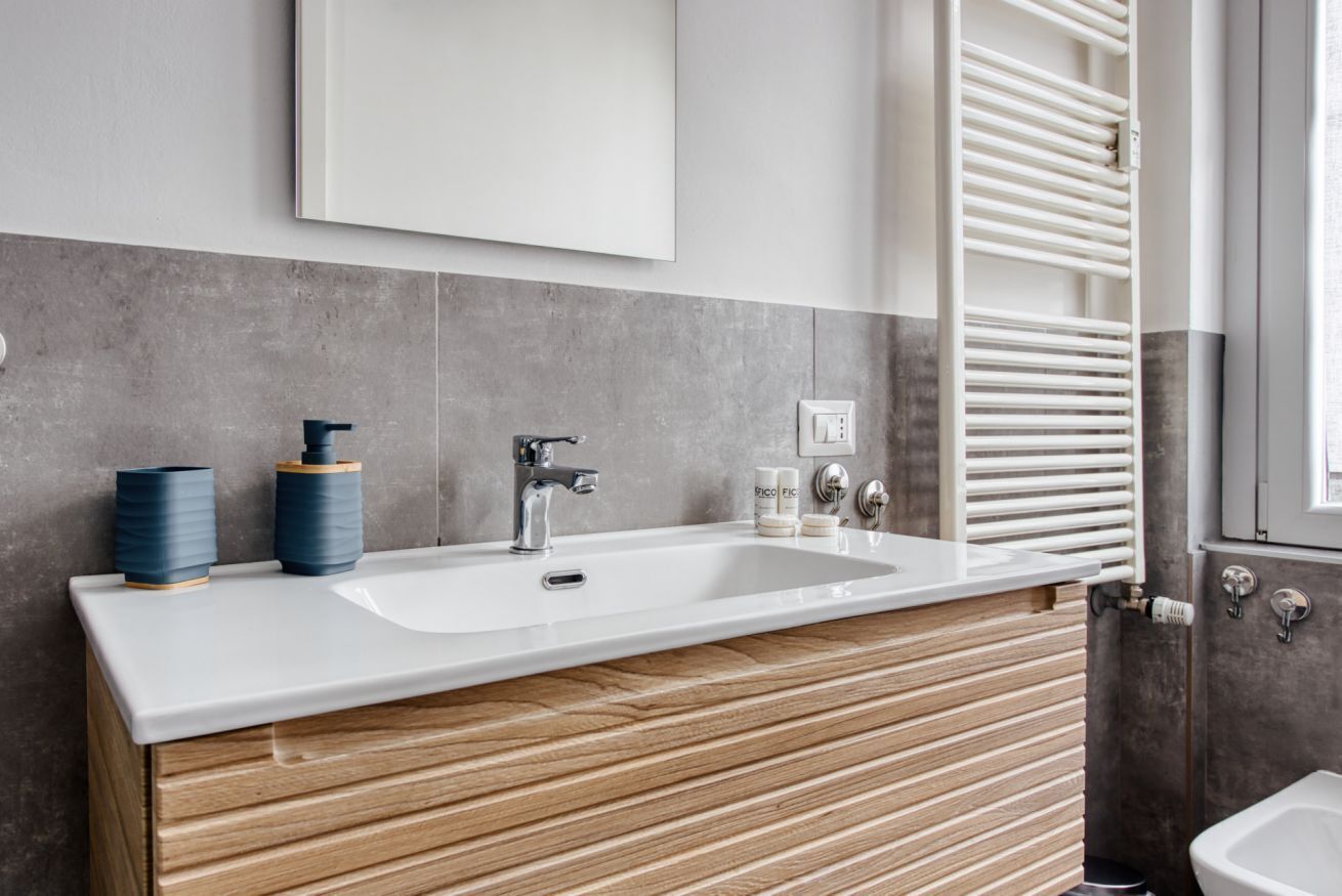 Un bagno moderno con un lavandino bianco, un mobile contenitore in legno, piastrelle grigie e un portasciugamani bianco.