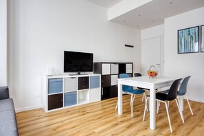 Un soggiorno moderno con una TV a schermo piatto, un tavolo da pranzo bianco con sedie blu, un divano grigio e un pavimento in legno.