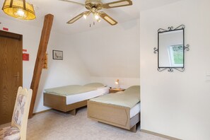 Une chambre avec deux lits simples, un ventilateur de plafond, une lampe de chevet, une chaise en bois et un miroir au mur.