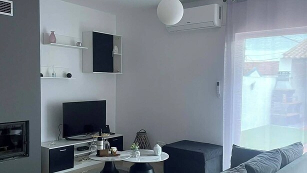A modern living room with a grey sofa, a coffee table, and a flat-screen TV. There is a window with curtains, and a ceiling-mounted air conditioning unit.