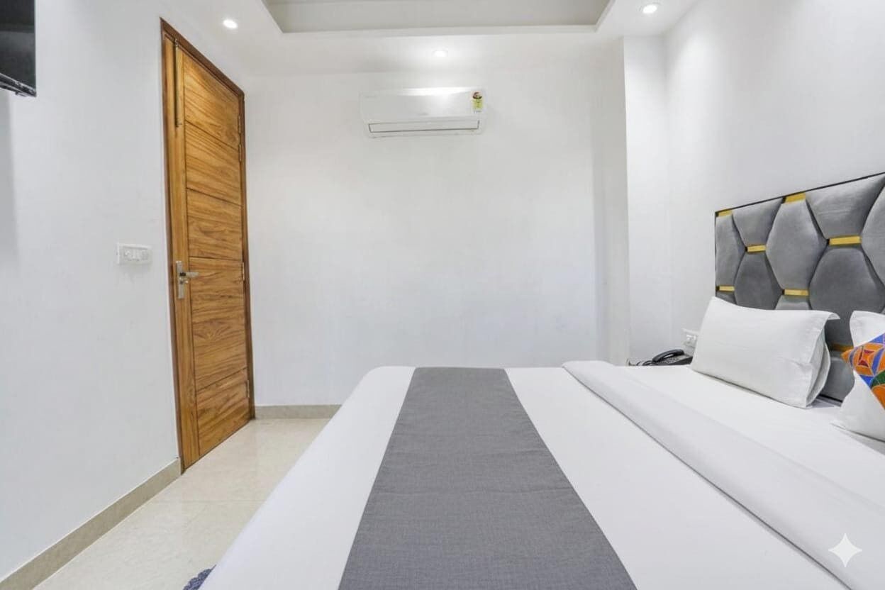 A bedroom with a wooden door, a bed with a grey headboard, and a small air conditioning unit on the wall.