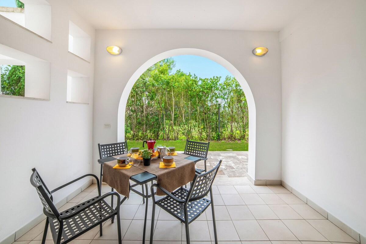 A dining area with a table and chairs, a view of greenery through an archway, and a tiled floor.