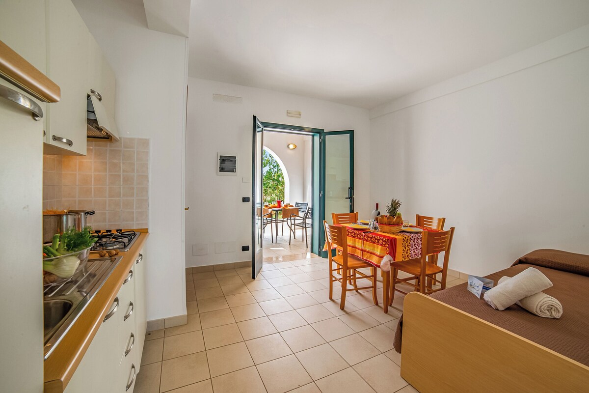 A compact hotel room with a kitchenette, a dining area with a table and chairs, and a bed with a towel.