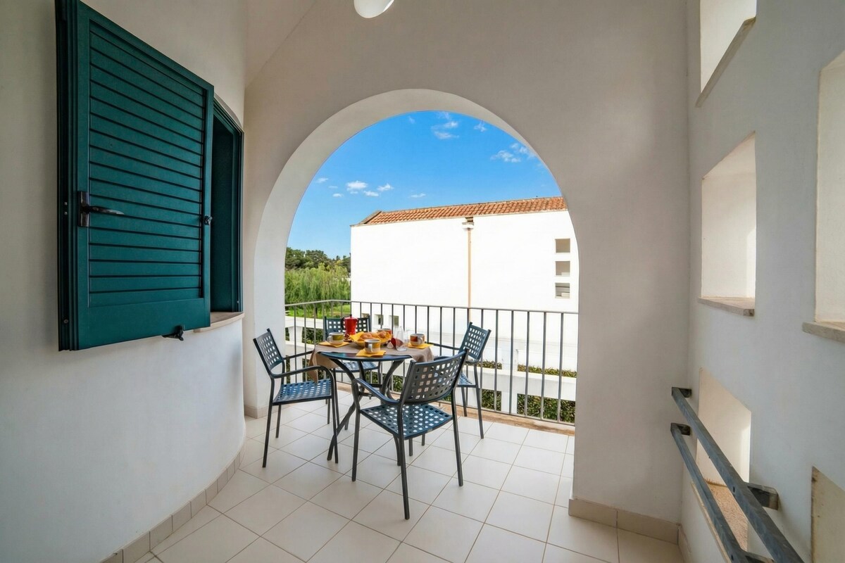 A balcony with a table and chairs, a green shuttered window, and a view of a white building.