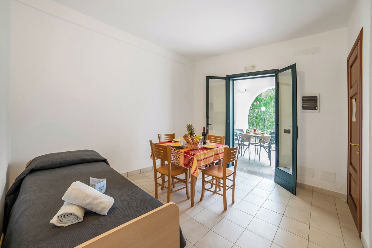 A bedroom with a bed, a dining table with chairs, and a view through an open door.
