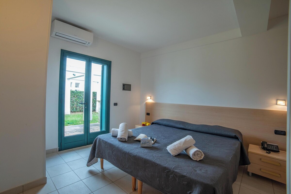 A hotel room with a bed, a bedside table with a phone, a nightstand, a window with a view, and an air conditioning unit.