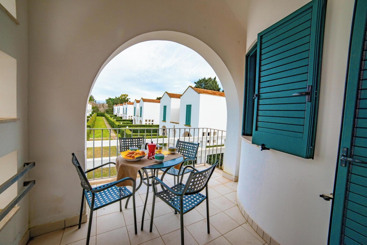 A balcony with a table and chairs, a view of white buildings with green shutters, and a tiled floor.