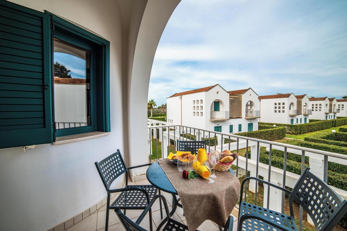 A balcony with a table set for two, a window with green shutters, and a view of white buildings with red roofs.
