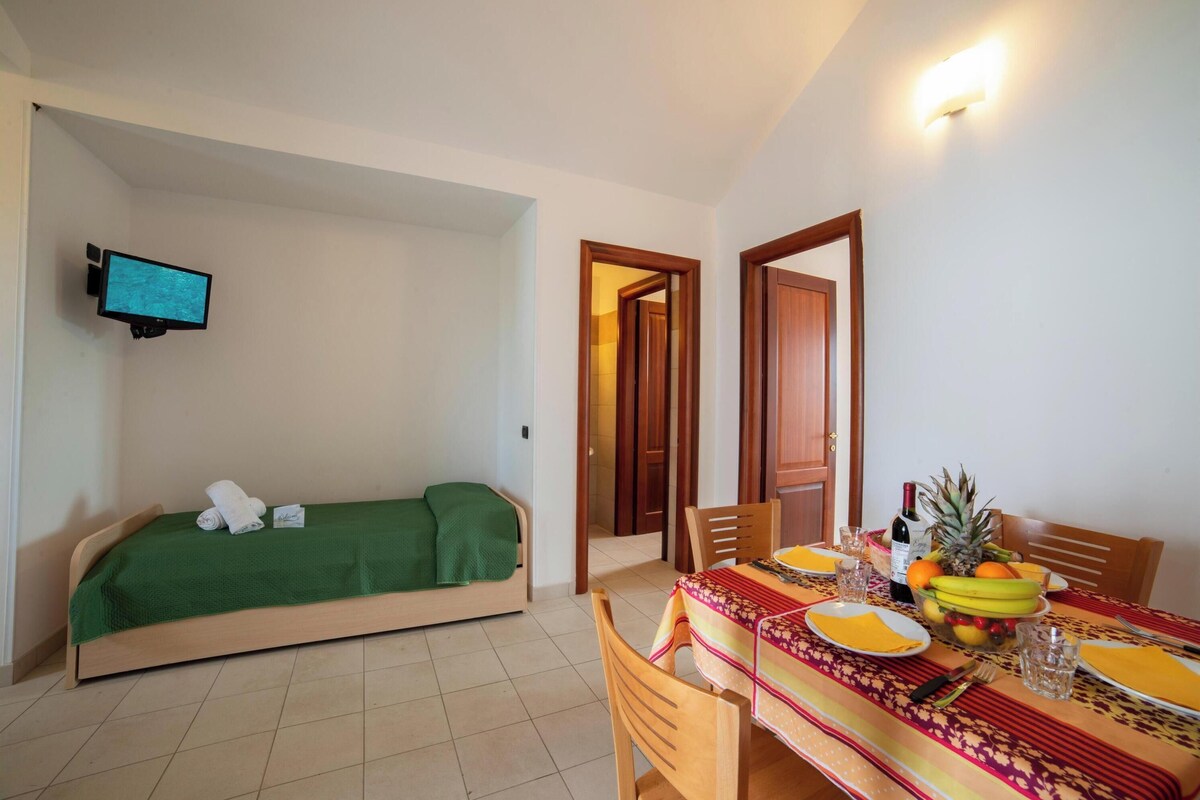 A room with a bed, a dining table set with plates, glasses, and a fruit bowl.