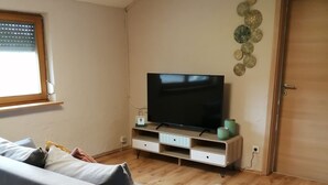 Een woonkamer met een grijze bank, houten vloeren, een flatscreen televisie op een standaard, en een deur aan de rechterkant.